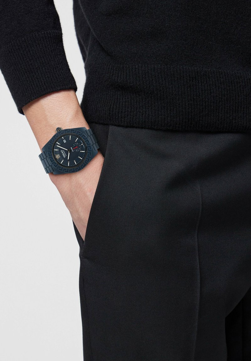 Linkerhand in zwarte broekzak, draagt een zwarte trui en een donkere horloge met een gepolijst bezel en een zichtbare datum op de wijzerplaat.
