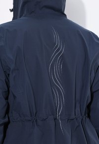 Marinblå vattentät jacka bakifrån med cinchad midja och vertikal reflekterande prickad vågmönsterdetalj, bärs av en person som står med ryggen mot kameran.