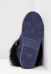 Pantufla azul marino con suela de goma texturizada y acentos de piel sintética. Material de ante suave, forma curva, con etiqueta de tamaño 37 en el lado.
