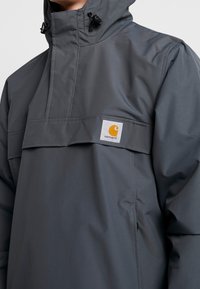 Grå vattentät pullover-jacka med hög krage, justerbara snoddar och en framficka med en logotypapplikation. Slen textur, funktionell design.