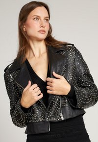 Giacca cropped in pelle nera con rivetti argentati, chiusura con zip, rever appuntiti e dettagli testurizzati sulle maniche. Senza ulteriori decorazioni.