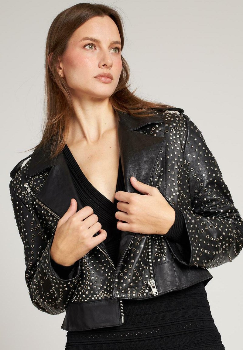 Giacca cropped in pelle nera con rivetti argentati, chiusura con zip, rever appuntiti e dettagli testurizzati sulle maniche. Senza ulteriori decorazioni.