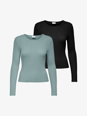 Kvinde med langt brunt hår iført en sort langærmet top, høj talje, vidde blå jeans og sorte sko, stående foran en ensfarvet baggrund.