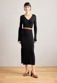 Black ribbed long-sleeve dress with a V-neck and button details, cinched with a tan leather belt; brown heeled shoes complete the outfit.