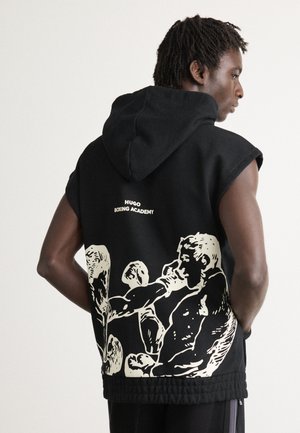 Uomo con un hoodie senza maniche di colore nero con la scritta "Hugo Boxing Academy" e una grafica con due pugili sulla schiena, rivolto di lato con le mani nelle tasche.