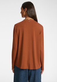 Top a maniche lunghe color ruggine realizzato in tessuto morbido. Presenta un collo rotondo e un leggero design con orlo asimmetrico.