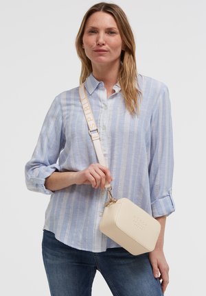 Woman wearing light blue striped button-up shirt and dark jeans, holding beige rectangular crossbody bag with wide strap.