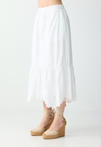 Femme portant une jupe midi blanche à ourlet festonné et des espadrilles compensées beiges avec des brides autour de la cheville, debout sur un sol blanc uni.
