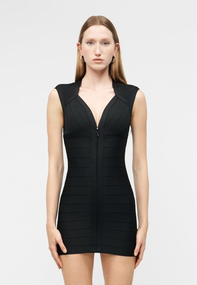 Femme aux cheveux longs portant une mini-robe noire ajustée sans manches avec un décolleté en V profond et une fermeture éclair à l'avant, debout devant un fond clair uni.