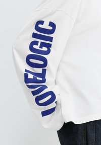 White long-sleeve shirt sleeve with bold blue text "LOVELOGIC" printed vertically, paired with dark denim jeans.