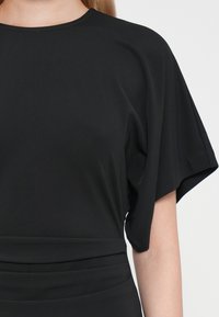 Crni kratki rukav majica napravljena od mekanog materijala, s okruglim izrezom i detaljem uskog struka za uskoj siluetu.