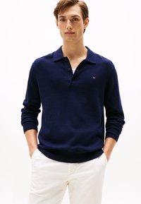 Suéter de polo de punto azul marino con una tapeta de tres botones y puños de canalé. Logo de acento blanco en el pecho. Llevado con pantalones de color claro.