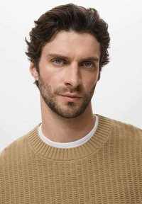 Beige gebreide trui met een geribbelde structuur, met een ronde halslijn en over een wit shirt gedragen. Effen achtergrond.