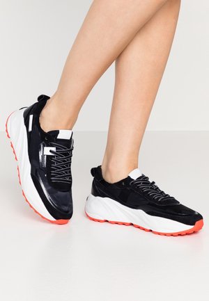 Sneaker chunky bianche e nere con suole rosse indossate su gambe nude incrociate contro uno sfondo uniforme.