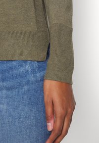 Olivenfarbener Strickpullover mit gerippten Bündchen, kombiniert mit blauen Jeans. Glatte Textur mit einer taillierten Passform um die Handgelenke.