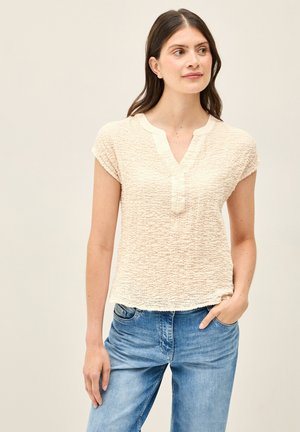 Korte mouwen, textuur crèmekleurige top met een V-hals en knoopdetail, gecombineerd met lichtblauwe jeans. Zacht materiaal en een relaxte pasvorm.
