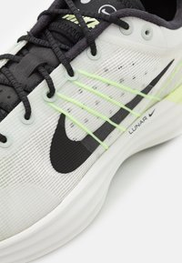 Šedá a černá sportovní obuv s pleteným svrškem, doplněná neonově zelenými detaily, černým logem swoosh a texturovanými tkaničkami na bílé podrážce.