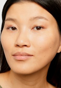 Gros plan sur un visage avec une peau lisse, un maquillage subtil mettant en valeur les yeux, et des cheveux noirs raides. Couleur des lèvres naturelle, fond neutre.