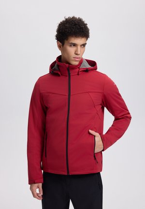 Mann trägt eine rote Kapuzenjacke mit Reißverschluss, hat die Hand in der Tasche und schaut seitlich vor einem schlichten hellen Hintergrund.