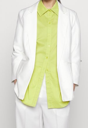 Persoon draagt een fel limoengroen overhemd met knopen onder een witte blazer en bijpassende witte broek, handen in de zakken van de blazer.