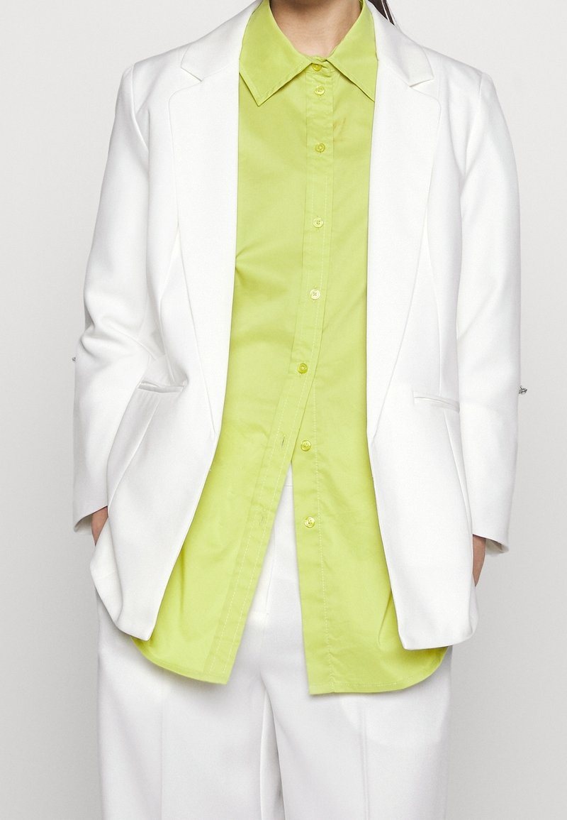 Personne portant une chemise boutonnée vert citron vif sous un blazer blanc et un pantalon blanc assorti, les mains dans les poches du blazer.