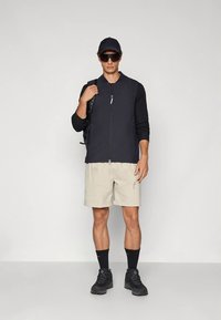 Juoda striukė su užtrauktuku, smėlio spalvos šortai, juodos kojinės ir batai. Aksesuarai: juoda kepurė ir akiniai nuo saulės. Kuprinė užmesta ant dešinio peties.