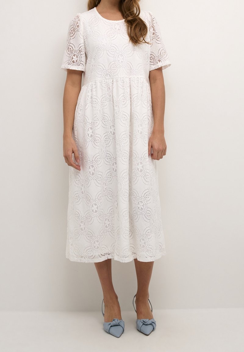 Femme portant une robe midi blanche en dentelle à manches courtes et des chaussures bleu pointues avec des nœuds, debout devant un fond uni.