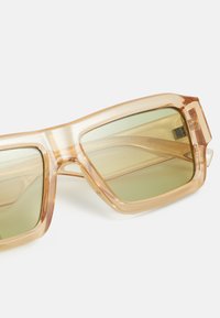 Transparent light brown oversized sunglasses with green tinted lenses, angular shape, and thick frames. Note the subtle texture and sleek design.