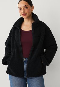 Zwarte fleecjas met een hoge kraag, ritssluiting en een zachte textuur. Draag het over een bordeauxrood topje en combineer het met blauwe jeans.