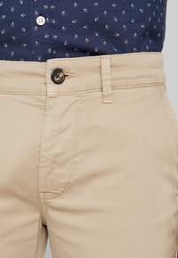 Béžové bavlněné chino kalhoty s klasickým rovnými nohavicemi, zapínáním na knoflík a hladkým materiálem s viditelnými detaily švů.