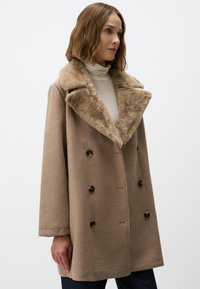 Cappotto in misto lana marrone con colletto in ecopelliccia, design doppiopetto e grandi bottoni marroni. Presenta maniche lunghe e una vestibilità comoda.