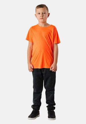 Ragazzo in piedi, volto rivolto verso avanti, che indossa una maglietta a maniche corte di un arancione brillante, pantaloni della tuta neri e scarpe da ginnastica nere su sfondo bianco.