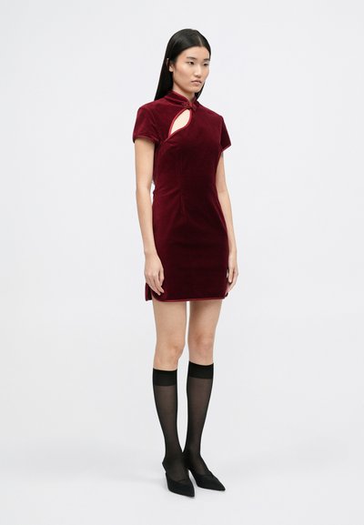 Rochie din catifea bordeaux, cu guler înalt, mâneci scurte și decupaj pe lateral, având detalii decorative cu nasturi. Modelul poartă ciorapi negri până la genunchi.