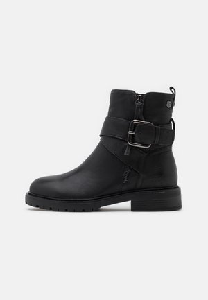 Bottine en cuir noir avec un bout arrondi, fermeture éclair sur le côté et une sangle décorative avec boucle. Surface texturée et talon épais.
