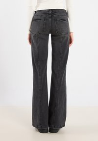 Pantalones vaqueros de mezclilla negra acampanados con cintura alta, dos bolsillos traseros y costuras visibles. Textura suave con un ligero brillo.