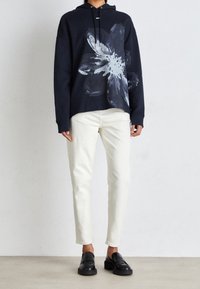 Mörkblå hoodie med stor vit blommig grafik, i kombination med off-white slim-fit byxor och svarta loafers mot en ljus bakgrund.