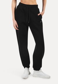 Henkilö, jolla on mustat jogging-housut joustavilla hihoilla ja valkoiset lenkkarit, yksi käsi taskussa, yhtenäistä taustaa vasten.