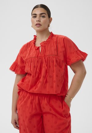 Woman wearing red eyelet lace top with ruffled sleeves and matching loose pants, standing with one hand in pocket on gray background.