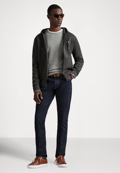 Γκρι φούτερ με κορδόνια, γκρι πουλόβερ, σκούρο τζιν και καφέ αθλητικά παπούτσια. Αξεσουάρ περιλαμβάνουν γυαλιά ηλίου και ρολόι.