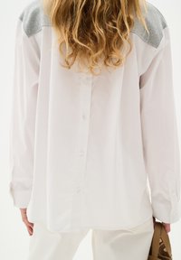 Camicia bianca a maniche lunghe con tessuto grigio sulle spalle, dotata di un retro con bottoni e una vestibilità ampia, abbinata a pantaloni beige chiaro.