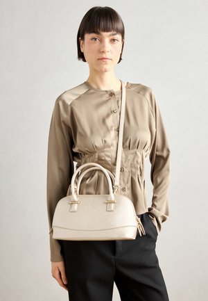 Femme aux cheveux courts et foncés portant un chemisier beige boutonné et un pantalon noir, tenant un sac à main beige avec une bandoulière.
