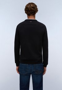 Homme aux cheveux courts et bouclés portant une chemise noire à manches longues et un jean bleu, de dos devant un fond uni clair.