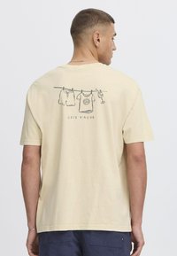 Lysegul bomulds-t-shirt med grafik af hængende tøj og en drink, med "CÔTE D'AZUR" trykt nedenunder. Korte ærmer og afslappet pasform.