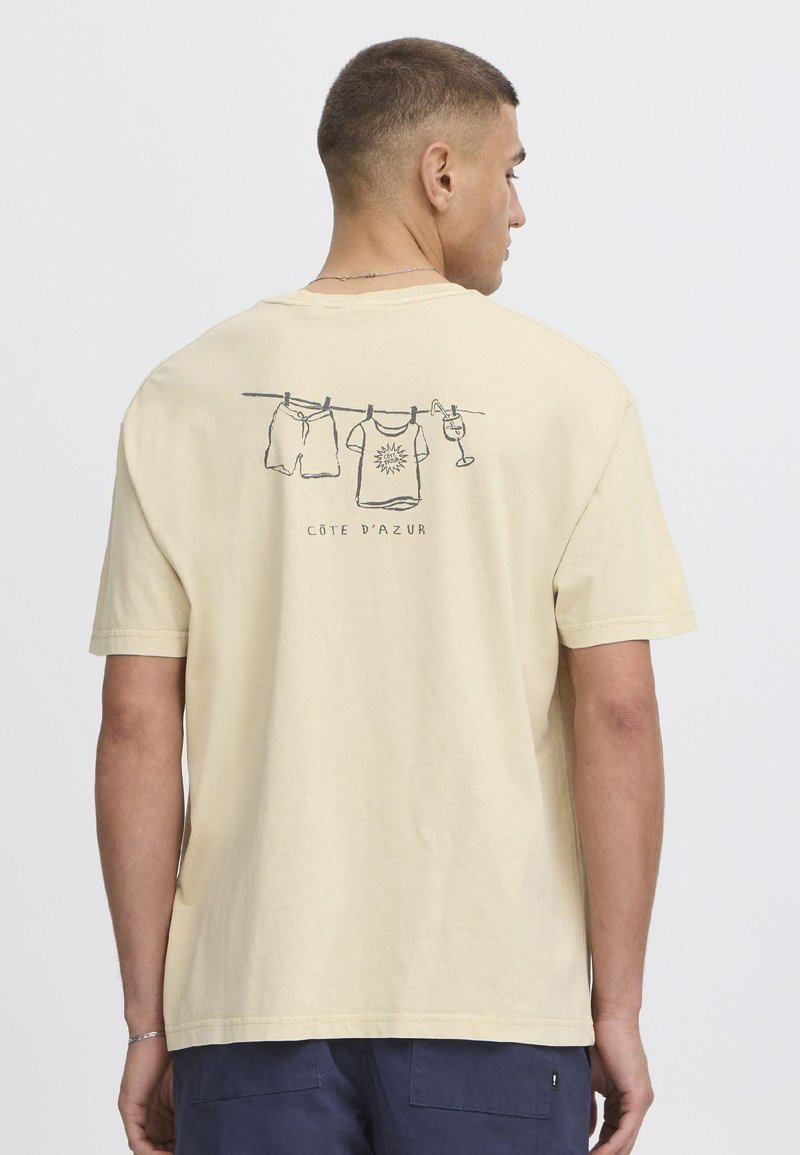 Lysegul bomulds-t-shirt med grafik af hængende tøj og en drink, med "CÔTE D'AZUR" trykt nedenunder. Korte ærmer og afslappet pasform.