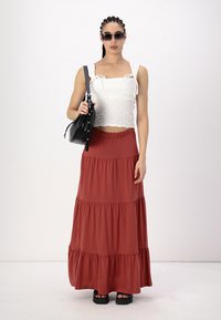 Biely smockovaný crop top s nastaviteľnými ramienkami, skombinovaný s rustikovo sfarbenou tiered maxi sukňou a čiernou kabelkou. Modelka nosí tmavé slnečné okuliare.