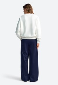 Balts džemperis ar vaļēju piegriezumu un ribotām aprocēm, kombinēts ar tumši zilām plaša piegriezuma biksēm. Mīksts audums, minimālistisks dizains, gluda tekstūra.