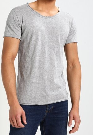 Mężczyzna w prostym, jasnoszarym t-shircie z krótkim rękawem i ciemnoniebieskich dżinsach, stojący na jednolitym tle.