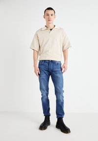 Polo de manga corta beige con collar blanco, combinado con jeans de mezclilla azules. El modelo lleva botas negras hasta el tobillo con detalles en la suela amarilla.