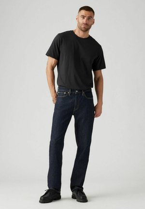 Black short-sleeve t-shirt paired with dark blue straight-leg jeans. Classic denim design with brass hardware and contrasting stitching. Black shoes.