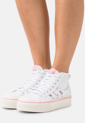 White canvas high-top sneakers with pink accents, featuring a textured rubber sole, floral design on the side, and white laces.